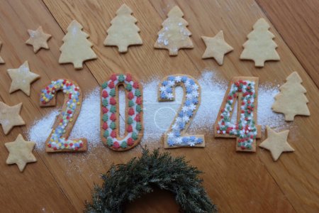 Foto de Galletas de corteza corta con espolvoreos de colores en forma de números 2024 con guirnaldas de pino y adornos en mesa de madera - Imagen libre de derechos