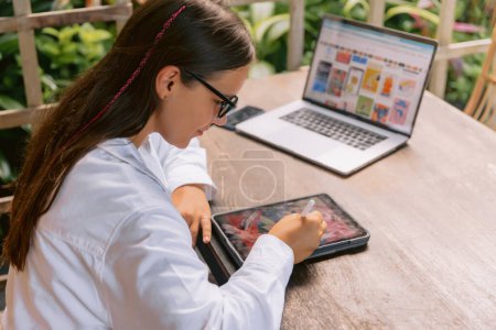 Téléchargez les photos : Attrayant jeune femme designer en utilisant tablette numérique et ordinateur portable dans le bureau d'été - en image libre de droit