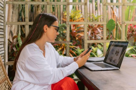 Téléchargez les photos : Attrayant jeune femme en utilisant smartphone et ordinateur portable dans le bureau d'été - en image libre de droit