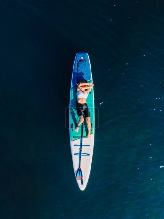 Téléchargez les photos : Young girl relaxing on stand up paddle board at quiet sea. Woman on SUP board in sea with morning light. Aerial view - en image libre de droit