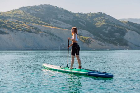 Téléchargez les photos : Young sporty woman rowing on stand up paddle board at quiet sea with beautiful landscape. - en image libre de droit