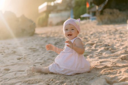 Téléchargez les photos : Portrait de bébé fille riante en robe rose sur la plage de l'océan avec la lumière du coucher du soleil. - en image libre de droit