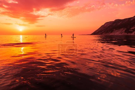 Photo for Travelers on stand up paddle board at quiet sea with bright sunset or sunrise. Girls on Red Paddle sup board and sunset with reflection - Royalty Free Image