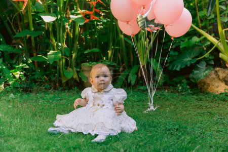 Téléchargez les photos : Anniversaire de mignon bébé fille avec des ballons à air dans le jardin extérieur. Joyeux enfant en robe. - en image libre de droit