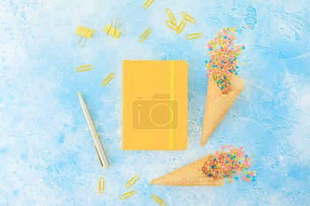 Téléchargez les photos : Journal avec stylo et bonbons aux confettis colorés et cône de gaufre sur bleu. Couché plat, vue du dessus - en image libre de droit