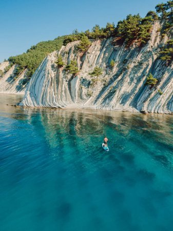 Téléchargez les photos : Homme debout planche à pagaie dans la mer bleue. Surfeur sur SUP board dans la mer Méditerranée avec paysage de montagne. Vue aérienne - en image libre de droit