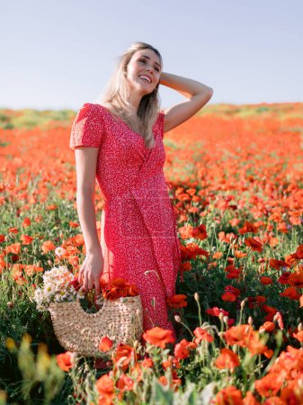 Foto de Atractiva mujer caucásica en un campo de amapola con flores. Mujer sonriente en vestido y bolso de paja con flores rojas - Imagen libre de derechos