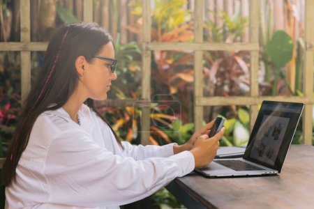 Téléchargez les photos : Femme pigiste utilisant un smartphone et un ordinateur portable dans un café d'été - en image libre de droit