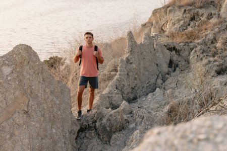 Photo for Handsome young man posing in a T-shirt and shorts. Hiker man stay on the coast among the rocks with warm sun light. - Royalty Free Image