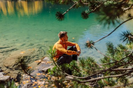 Foto de Hombre sentado cerca de un lago de cristal en las montañas otoñales. Lago transparente y relajante excursionista turista - Imagen libre de derechos