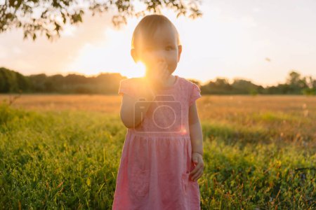 Téléchargez les photos : Enfant fille restant dans l'herbe avec des tons de coucher de soleil. Bébé mignon en robe rose - en image libre de droit