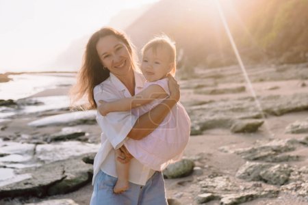 Téléchargez les photos : Mère joyeuse avec son bébé sur la plage avec du soleil. Happy famille en plein air - en image libre de droit