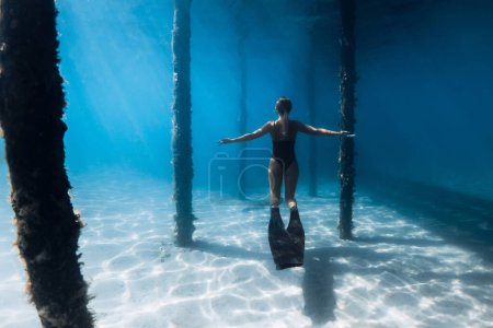 Téléchargez les photos : Femme plongeuse libre avec des nageoires posant sous la jetée dans l'océan bleu. Nage féminine avec des nageoires sous la jetée - en image libre de droit