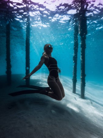 Mujer liberadora posando bajo el agua bajo el muelle en el océano azul. Mujer nada con aletas debajo del muelle