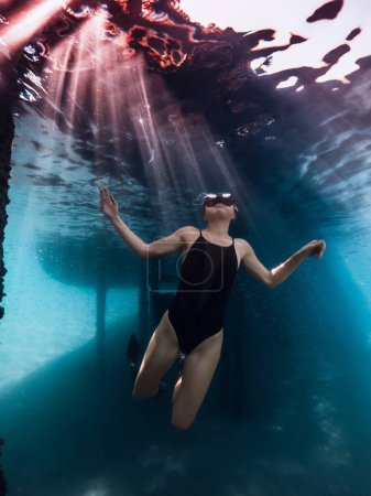 Téléchargez les photos : Femme plongeuse posant sous l'eau sous la jetée dans l'océan bleu avec des rayons de soleil. - en image libre de droit