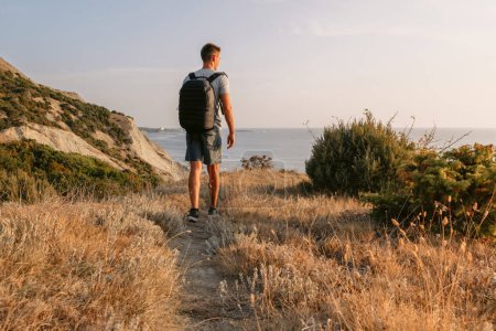 Téléchargez les photos : Beau randonneur avec sac à dos randonnée sur la côte de l'océan avec des montagnes et la lumière du soleil du soir. - en image libre de droit