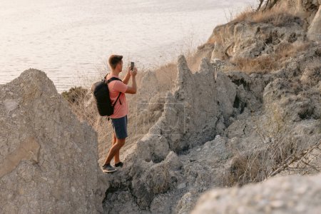 Téléchargez les photos : Randonneur homme restant à la roche sur la côte avec la lumière du coucher du soleil chaud. Voyageur homme avec smartphone en extérieur. - en image libre de droit