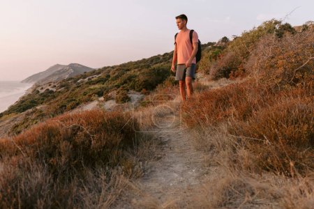 Téléchargez les photos : Randonneur homme avec sac à dos marchant sur la côte de l'océan avec la lumière du soleil chaud. - en image libre de droit