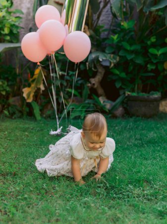 Téléchargez les photos : Anniversaire de mignon bébé fille avec des ballons à air dans le jardin extérieur. Bébé fille rampant sur l'herbe - en image libre de droit