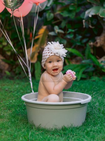 Photo for Happy baby girl and pink air balloons swimming in baby bath outdoor summer garden. - Royalty Free Image