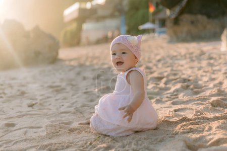 Photo for Happy smiling beautiful girl in outdoor on sandy beach with sunset light. Baby child in pink dress - Royalty Free Image