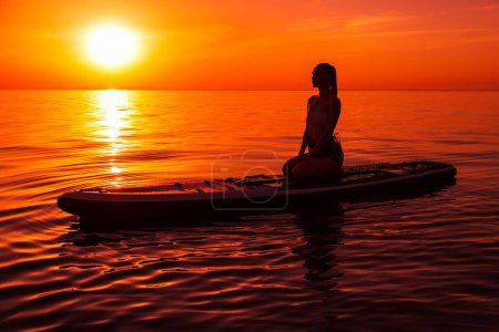 Téléchargez les photos : Silhouette de femme se relaxant sur planche à pagaie à la mer calme avec coucher de soleil lumineux chaud ou lever de soleil. - en image libre de droit
