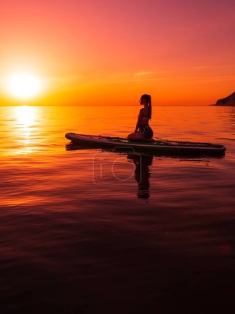 Téléchargez les photos : Femme mince profitant sur le stand up paddle board en mer avec coucher de soleil lumineux ou lever de soleil. Femme se détendre sur SUP board dans l'océan. - en image libre de droit