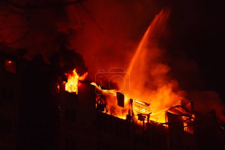 Foto de La casa ardiente está envuelta en llamas por la noche. Los bomberos se extinguen con agua. Incendio en edificio de apartamentos. - Imagen libre de derechos