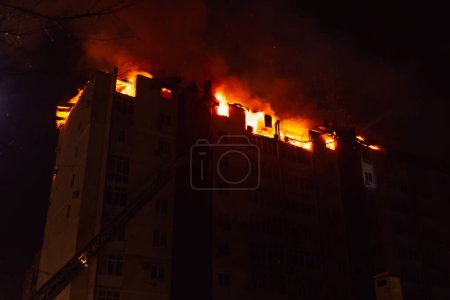 Téléchargez les photos : Énorme incendie flamboyant dans un immeuble à appartements de plusieurs étages. La maison en feu est engloutie dans les flammes la nuit pendant le désastre - en image libre de droit