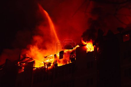 Photo for Burning house is engulfed in flames at night. Fire blazing in apartment building. Firefighters extinguish with water. - Royalty Free Image
