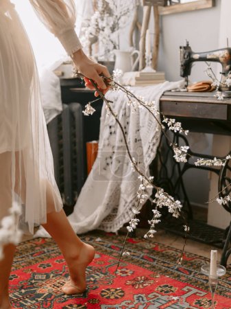 Téléchargez les photos : Jambes de femme à la mode portant une robe de mariée vintage posant à la maison dans un intérieur vintage élégant et tenant dans les mains branche fleurissante - en image libre de droit