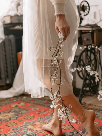 Foto de Las piernas de la mujer de moda con vestido de novia vintage posando en casa en el interior de la vendimia con estilo y celebrar en las manos rama de floración - Imagen libre de derechos