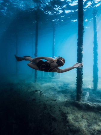 Free diver girl glides with fins dive under the pier in blue ocean. Female swims underwater dives between the pier pillars