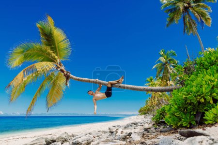 Téléchargez les photos : Homme torse nu relaxant sur la plage et grimpe un cocotier. Slim homme vacances sur la plage paradisiaque - en image libre de droit