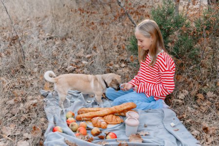 Téléchargez les photos : Young teenage girl with dog sitting on a plaid with drink and bakery on a picnic - en image libre de droit