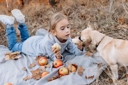 Téléchargez les photos : Jeune fille s'allonge sur un plaid, manger de la pomme et donner sandwich son chien sur un pique-nique - en image libre de droit