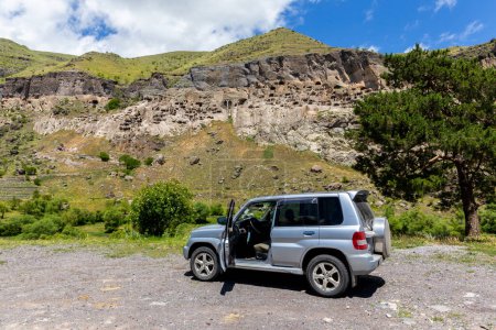 Photo for Off-road 4x4 car parked on a gravel road with Vardzia cave monastery complex in the background, Georgia. Erusheti Mountain with caves, tunnels and dwellings carved in the rock. - Royalty Free Image