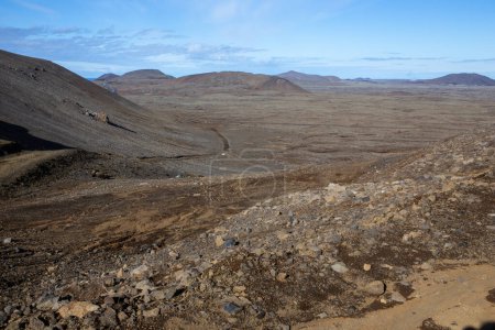 Barren Reykjanes peninsula landscape with old lava fields, dirt road and volcanic mountains in the background, blue sky.