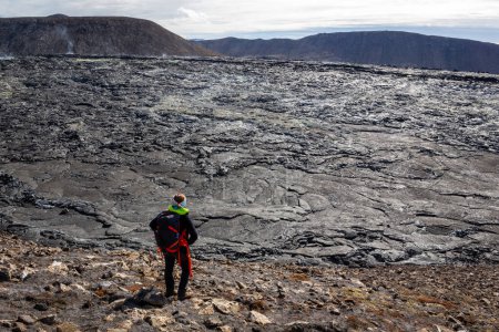Touristin mit Rucksack steht auf den geschmolzenen Basaltfelsen im Lavafeld des Vulkans Fagradalsfjall und beobachtet Vulkanlandschaft, Island.