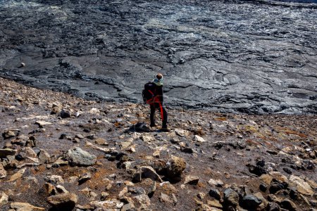 Female tourist with backpack standing on top of the molten basalt rocks in Fagradalsfjall volcano lava field, watching volcanic landscape, Iceland.