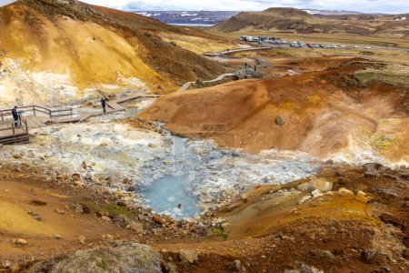 Seltun Geothermal Área en Krysuvik con aguas termales humeantes, colinas de azufre amarillo y naranja, personas caminando en paseos marítimos, aparcamiento en el fondo.