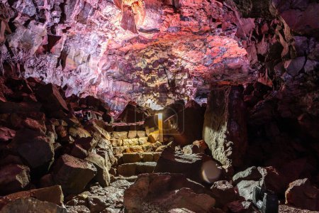 The Lava Tunnel (Raufarholshellir) in Iceland, inside view of the illuminated lava tube with red iron volcanic rock formations and stone walkway staircase for tourists.