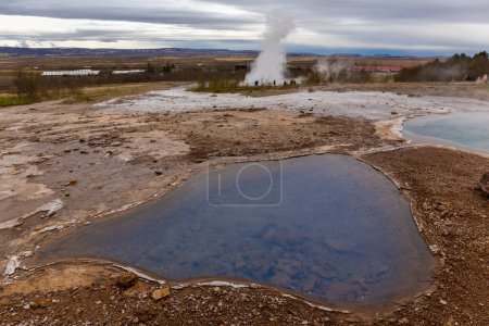 Geysir geothermal area landscape in Iceland with hot springs and pools, Strokkur geyser erupting in the background.