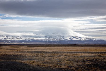 Raw Icelandic landscape with grasslands, lava fields and snowcapped Hekla volcano with peak covered in clouds.