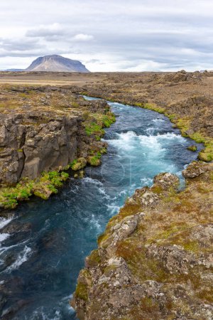 Thjorsardalur valley, South Iceland, landscape with turquoise glacial wild river, volcanic rock formations overgrown with moss and grass and mountains in the background.