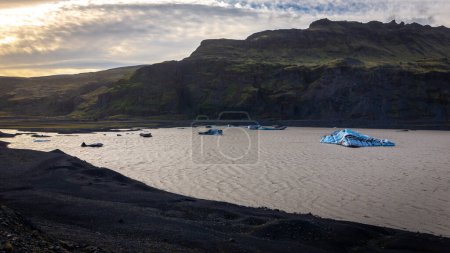 Solheimajokull, Teil des Myrdalsjokull-Gletschers, Gletscherlagune mit schwimmenden blauen Eisbergen und rauer Berglandschaft, Sonnenuntergangshimmel, Island.