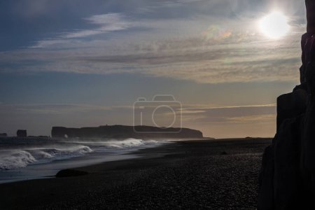Sneaker Wellen, die gegen die schwarze Sandstrandküste von Reynisfjara krachen, mit starkem Wind, der Wasser und vulkanischen schwarzen Sand bläst, Basaltsäulen im Vordergrund, Sonnenuntergang, Island.