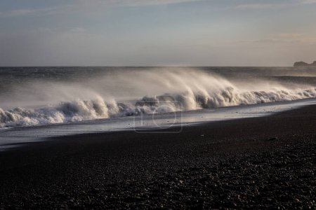 Sneaker-Wellen, die gegen die schwarze Sandstrandküste von Reynisfjara krachen, mit starkem Wind, der Wasser und vulkanischen schwarzen Sand bläst, Sonnenuntergang, extremes Wetter, Island.