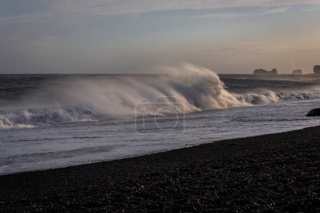 Sneaker olas aplastamiento contra Reynisfjara playa de arena negra costa, con fuertes vientos que soplan agua y arena negra volcánica, puesta de sol, clima extremo, Islandia.