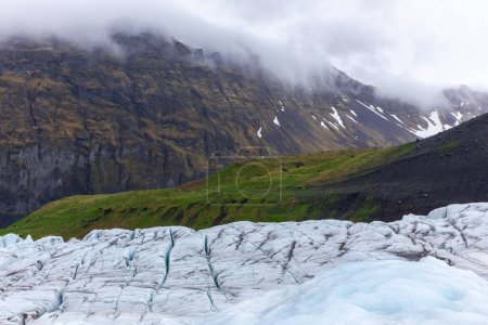 Mountain landscape with retreating Skaftafell Glacier in Vatnajokull National Park, Iceland. Blue glacier ice with cracks and crevasses, green grasslands and foggy mountain ridge in the background.